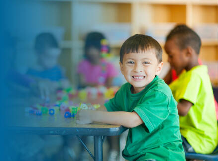 A smiling student in a green shirt sits at a classroom table while other students work with colorful learning materials in the background.